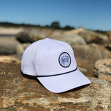 White baseball cap with round Blue Crab logo patch, resting on rocks at a sandy beach.