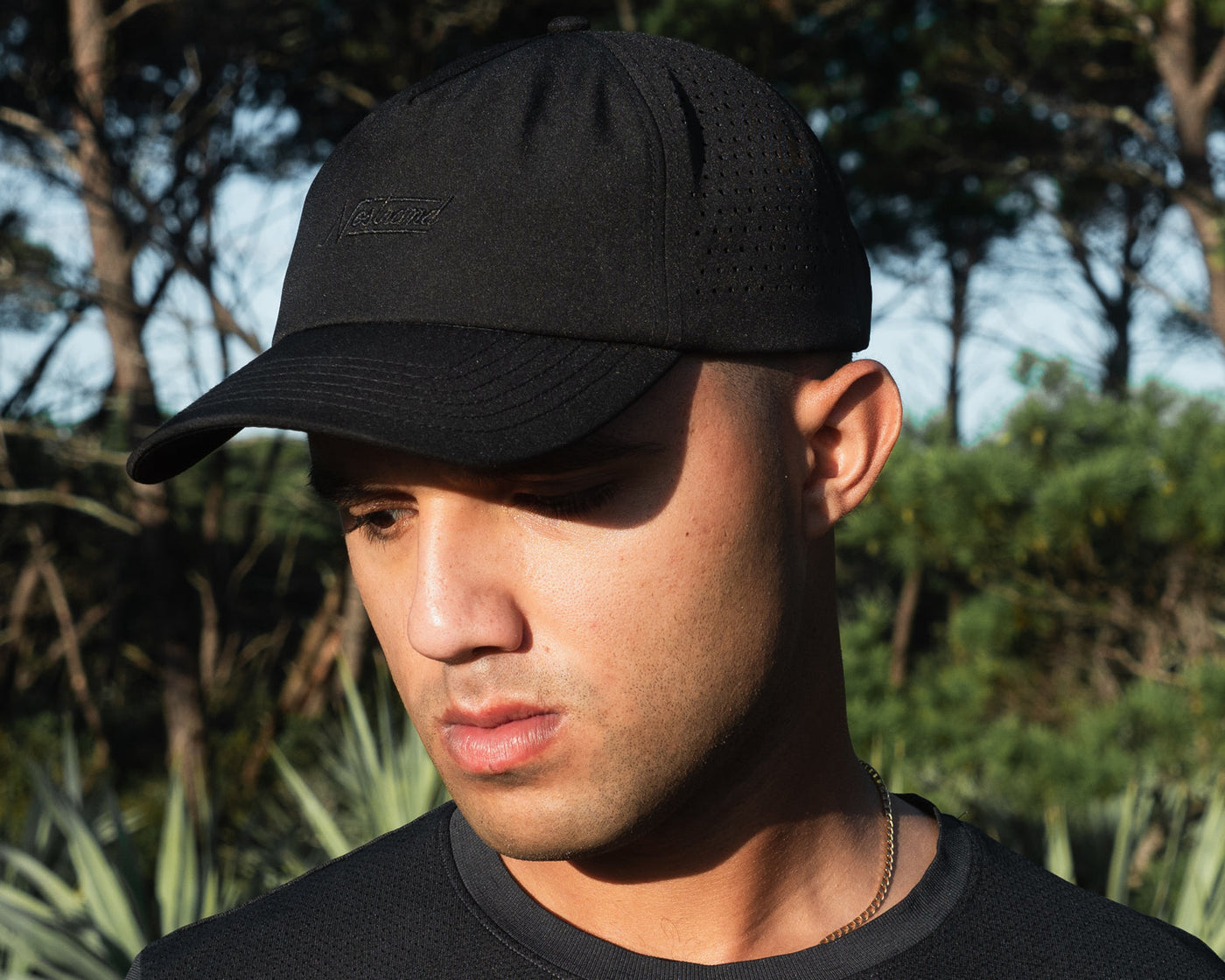 Three-quarter view of black golf hat on model with embroidered Nostrand logo and perforated side panels, trees behind