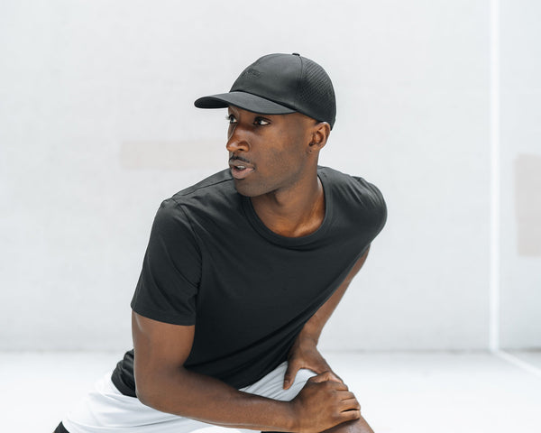 Person wearing a ventilated black baseball cap and black T-shirt, leaning forward and looking left - Agile Hat Black