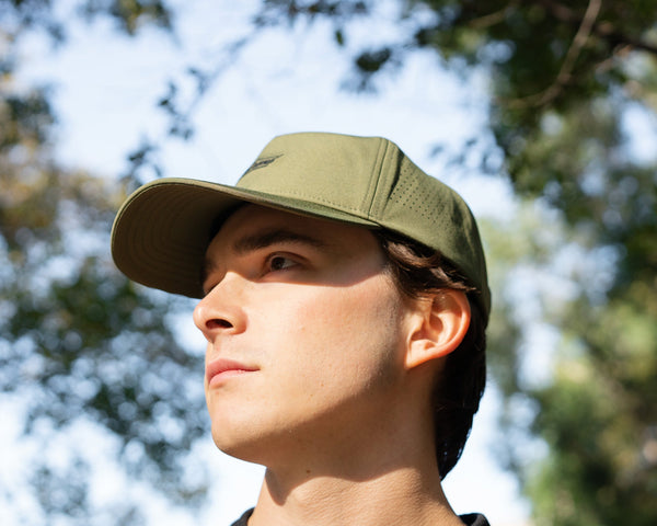 Young man in olive-green baseball cap looking upward, outdoor background with sunlit trees blurred - Daily Tech Hat Olive