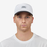A man facing forward wearing a white baseball cap with a black brim and a small rectangular 'Nostrand Sports' logo patch on the front, paired with a white t-shirt against a light gray background.