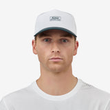 A man facing forward wearing a white baseball cap with a dark green brim and a small rectangular 'Nostrand' logo patch on the front, paired with a white t-shirt against a light gray background.