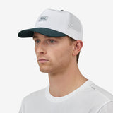 A man angled wearing a white baseball cap with a dark green brim and a small rectangular 'Nostrand' logo patch on the front, paired with a white t-shirt against a light gray background.