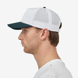 A man profiled wearing a white baseball cap with a dark green brim and a small rectangular 'Nostrand Sports' logo patch on the front, paired with a white t-shirt against a light gray background.