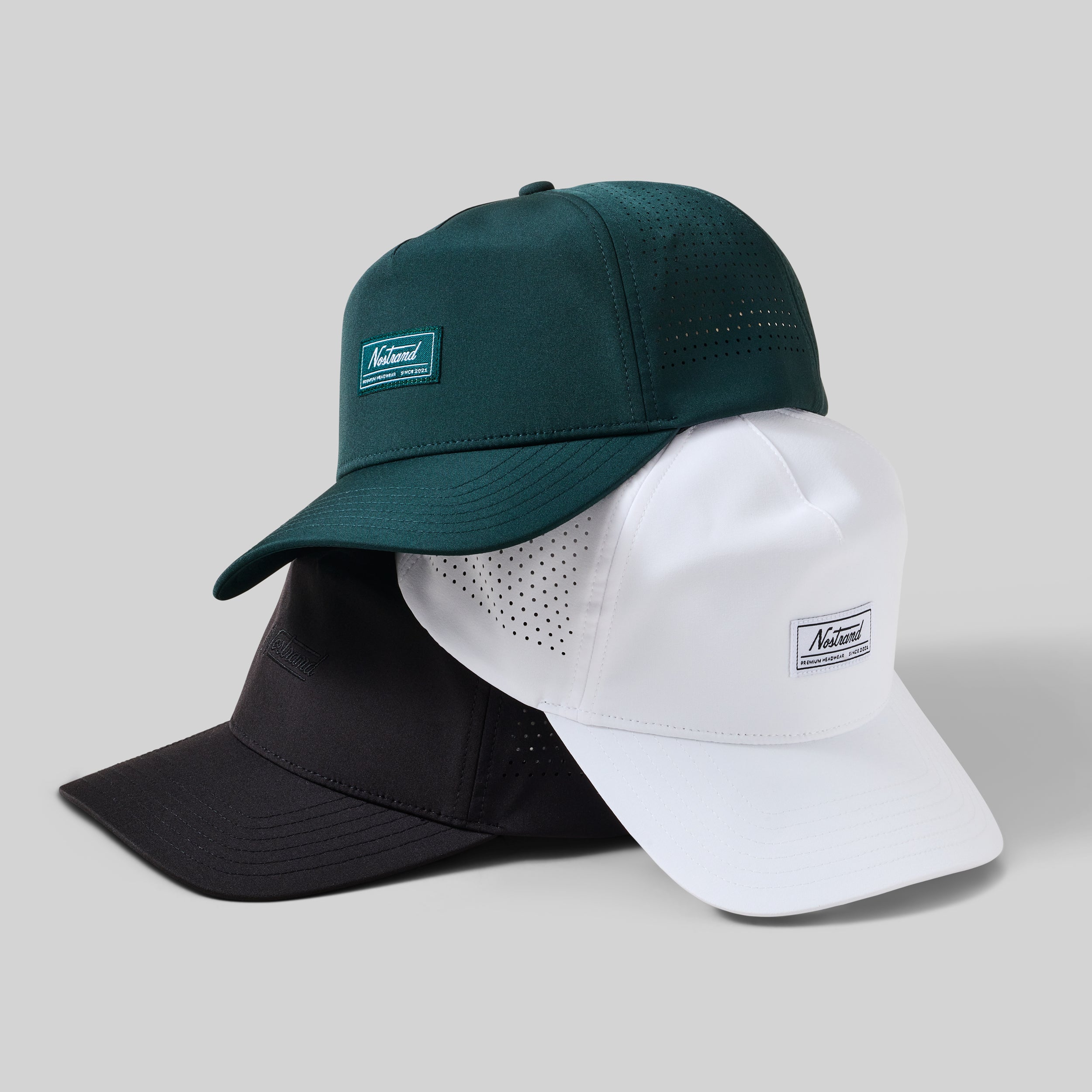 Three stacked performance caps in green, white, and black with a small rectangular Nostrand label on the front