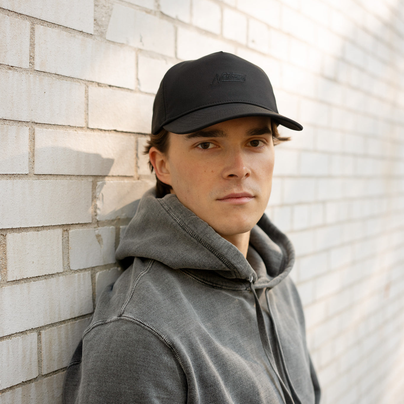Male model wearing black Daily Tech Hat 3 cap and gray hoodie, leaning against a light brick wall