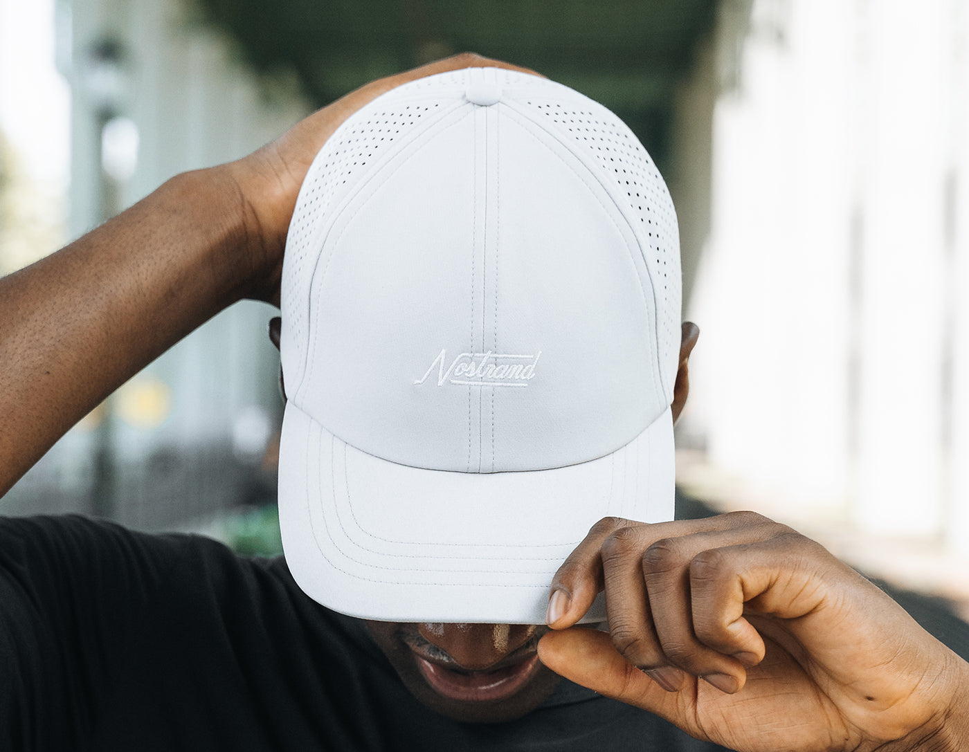 Person lowering white baseball cap embroidered with Nostrand, hand on brim, face mostly hidden - Agile Hat Grey